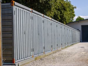 Longue rangée de box de stockage individuels gris numérotés sur une base en béton, le long d'un chemin de gravier. Arbres et bâtiment en arrière-plan.
