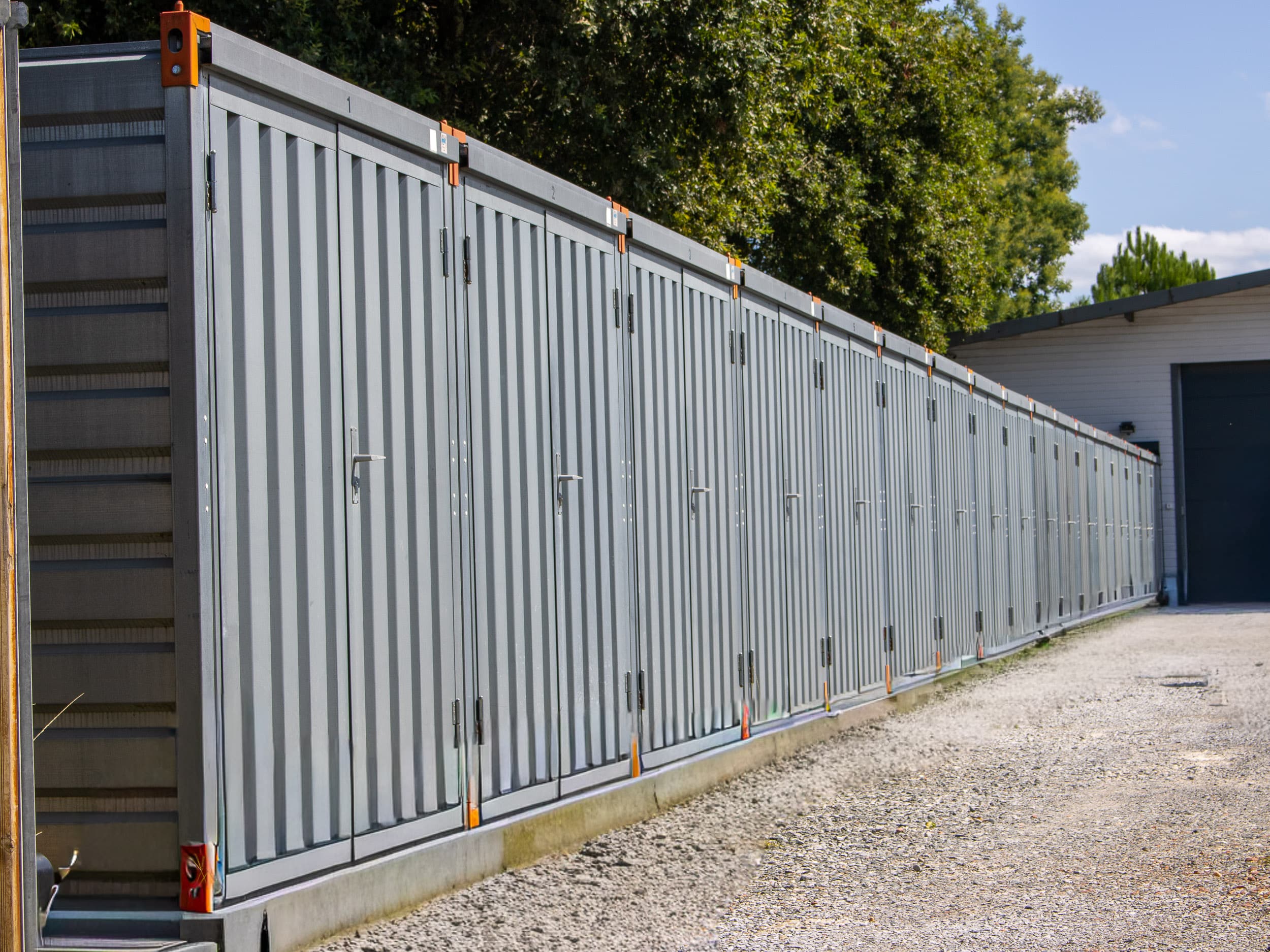 Longue rangée de box de stockage individuels gris numérotés sur une base en béton, le long d'un chemin de gravier. Arbres et bâtiment en arrière-plan.