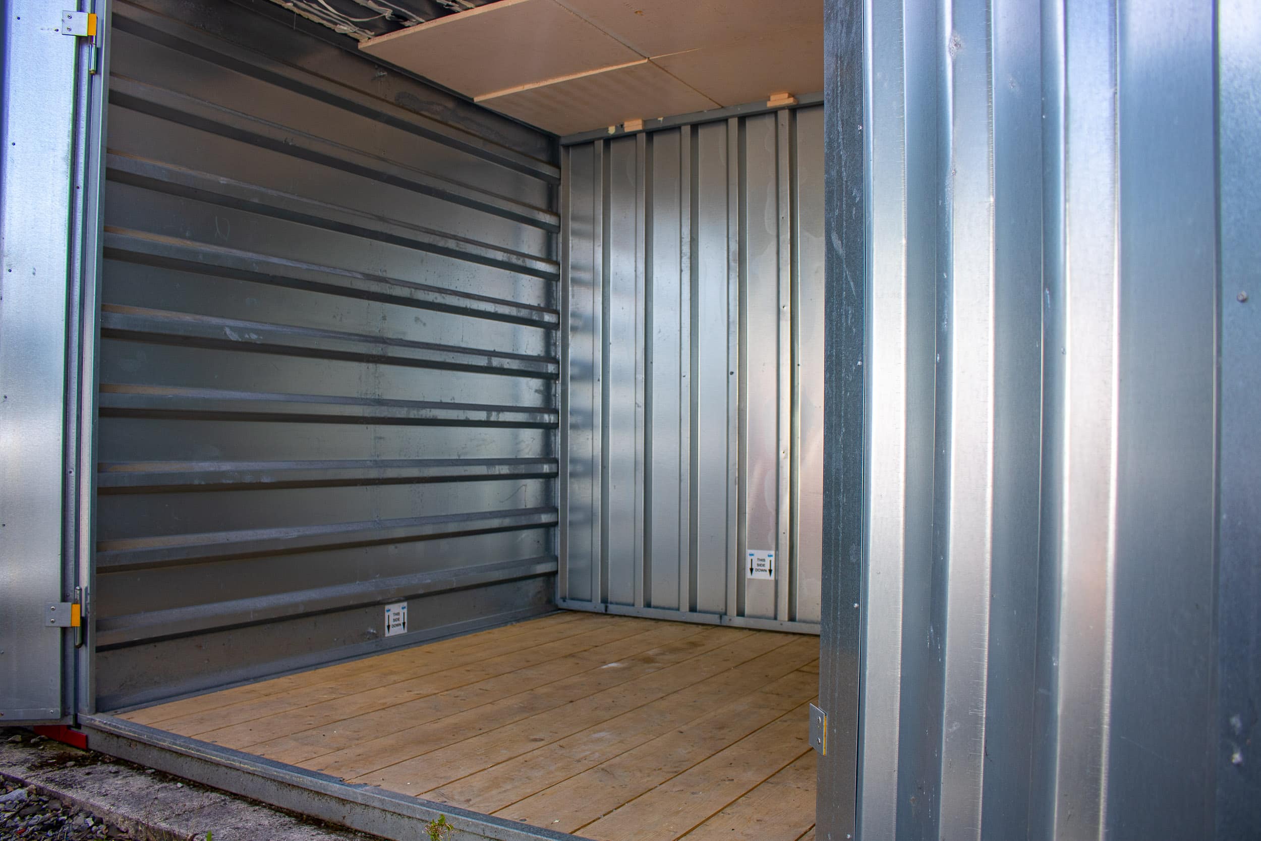 Intérieur d'un box de stockage vide en métal galvanisé et plancher bois. La porte ouverte révèle un autocollant "THIS SIDE DOWN".