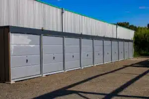 Rangée de boxes de stockage individuels gris numérotés 13 à 21, adossés à un entrepôt métallique sous un ciel bleu dégagé, sur sol gravier.