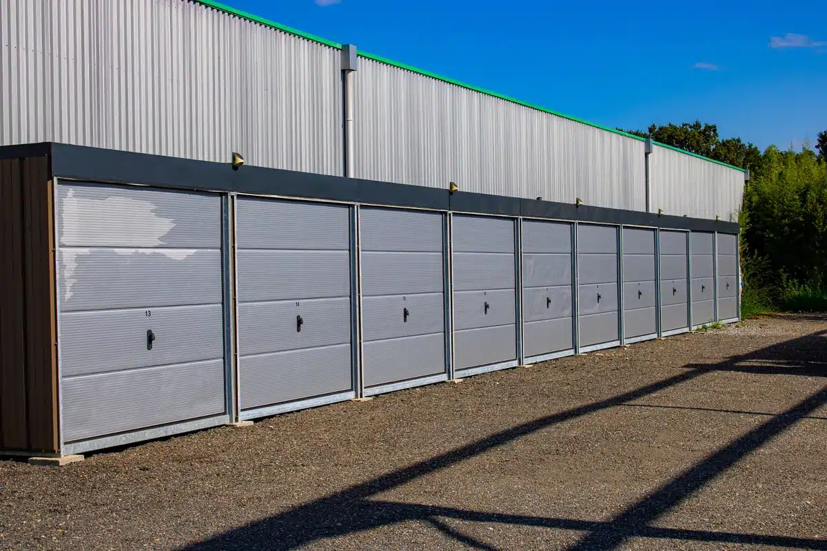 Rangée de boxes de stockage individuels gris numérotés 13 à 21, adossés à un entrepôt métallique sous un ciel bleu dégagé, sur sol gravier.