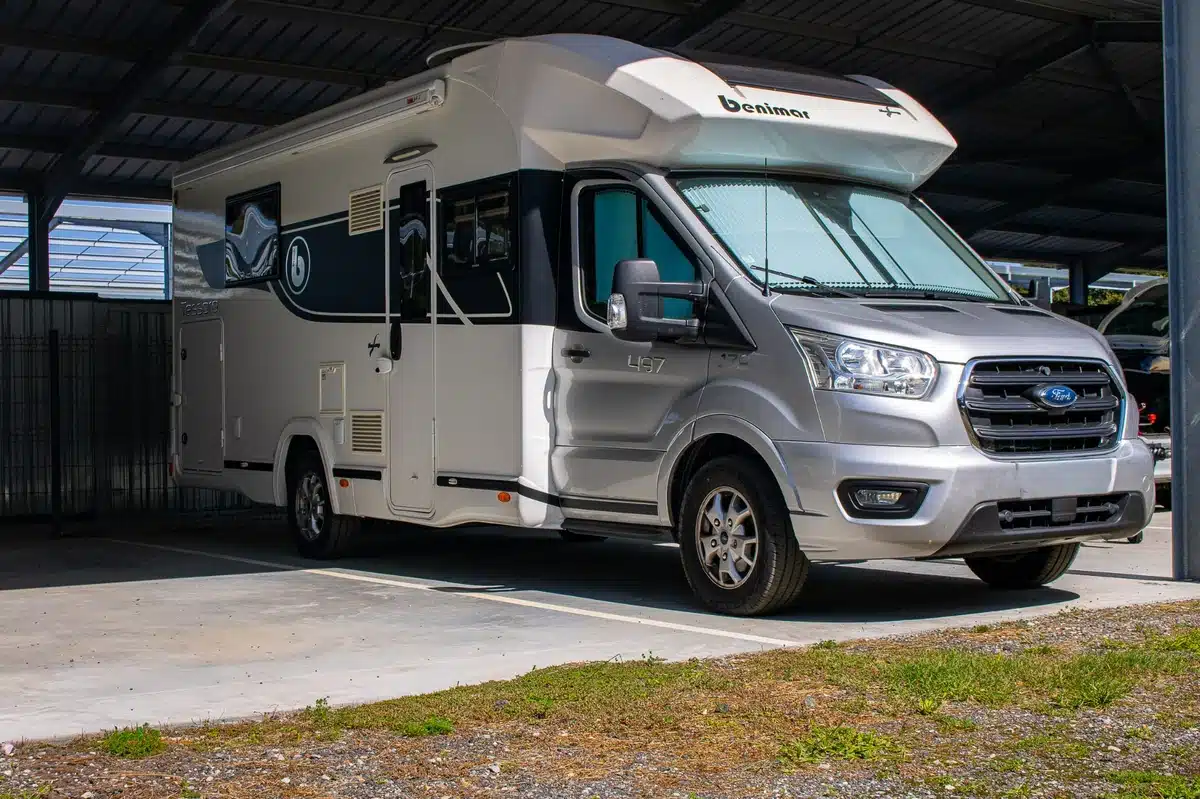 Camping-car Benimar 497 gris et blanc, basé sur châssis Ford, stationné sous un abri en métal.