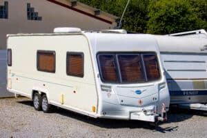 Caravane Bailey blanche à doubles essieux et stores, stationnée sur gravier à côté d'une caravane Fendt. Bâtiment avec pavés de verre. Texte: BAILEY, 63, FENDT.