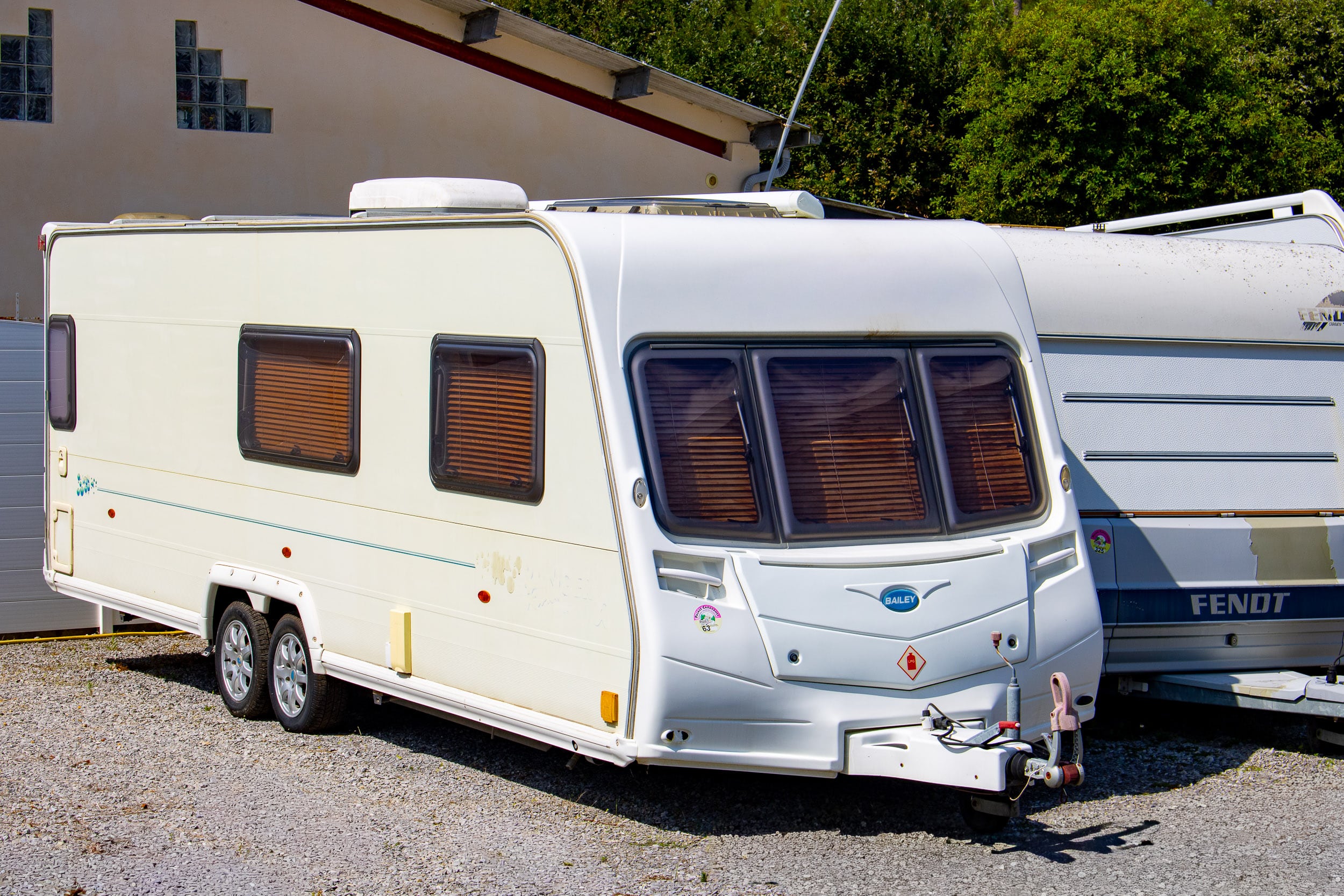 Caravane Bailey blanche à doubles essieux et stores, stationnée sur gravier à côté d'une caravane Fendt. Bâtiment avec pavés de verre. Texte: BAILEY, 63, FENDT.