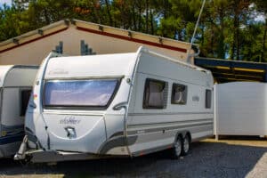 Grande caravane blanche Emotion Silver Dethleffs à double essieu, garée sur un sol en gravier devant un bâtiment crème et des arbres verts, avec son attelage visible à l'avant.