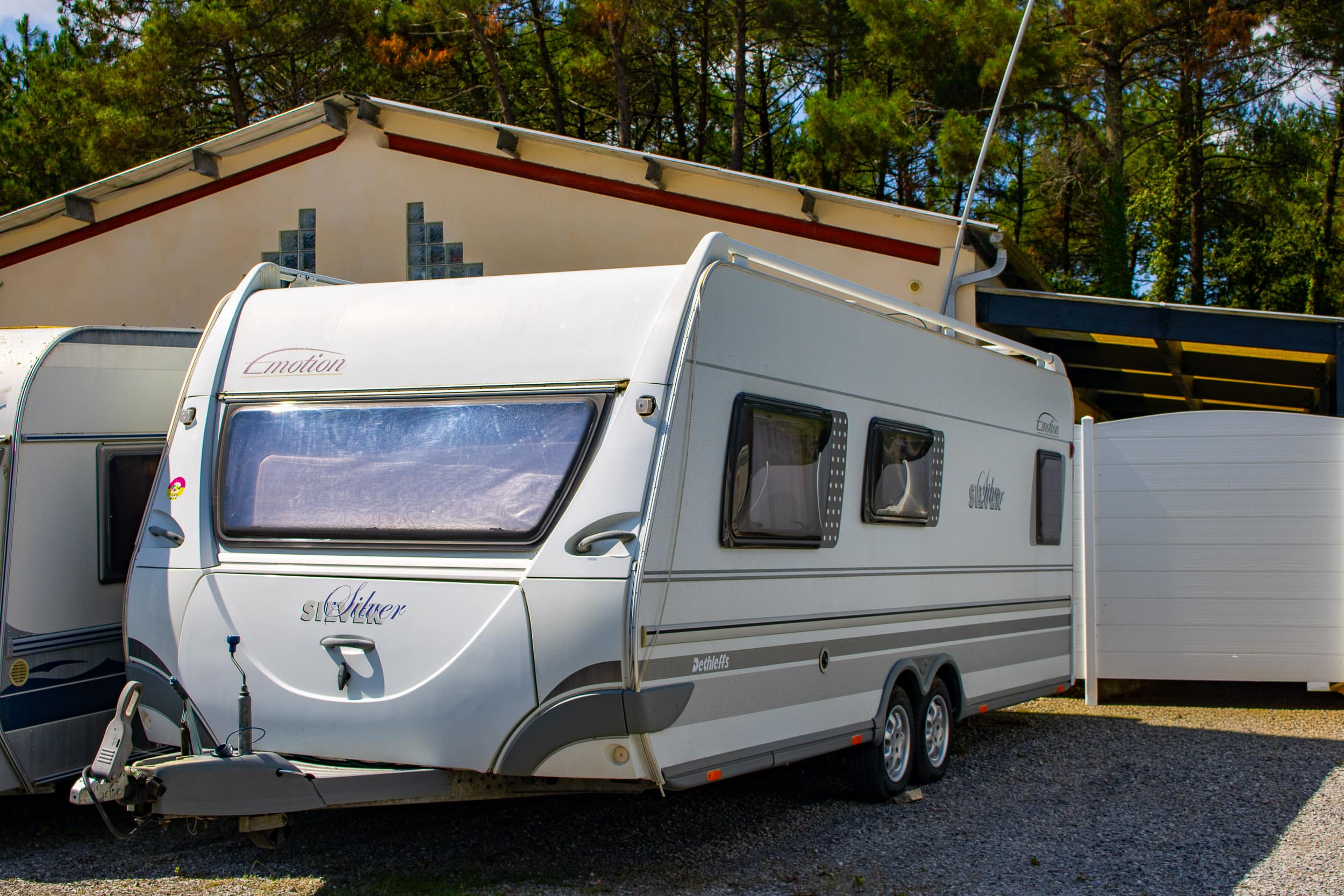 Grande caravane blanche Emotion Silver Dethleffs à double essieu, garée sur un sol en gravier devant un bâtiment crème et des arbres verts, avec son attelage visible à l'avant.