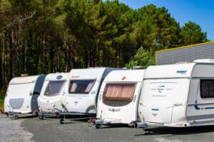 Rangée de caravanes Dethleffs, Burstner, Fendt, Caravelair, Avento, Bianco sur un parking, devant une forêt de pins et un bâtiment.