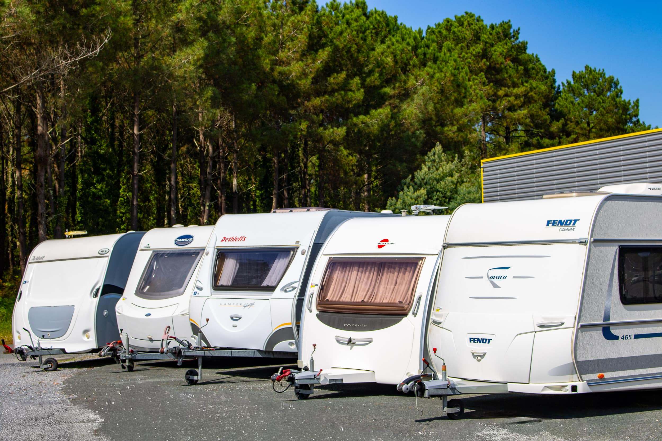 Rangée de caravanes Dethleffs, Burstner, Fendt, Caravelair, Avento, Bianco sur un parking, devant une forêt de pins et un bâtiment.
