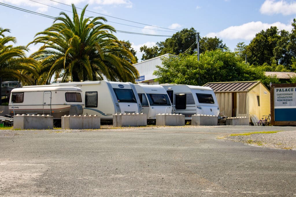 Caravanes garées sur un parking ensoleillé, bordé de palmiers. Panneau de services PALACE P: Gardiennage, Réparations, Remorquage.