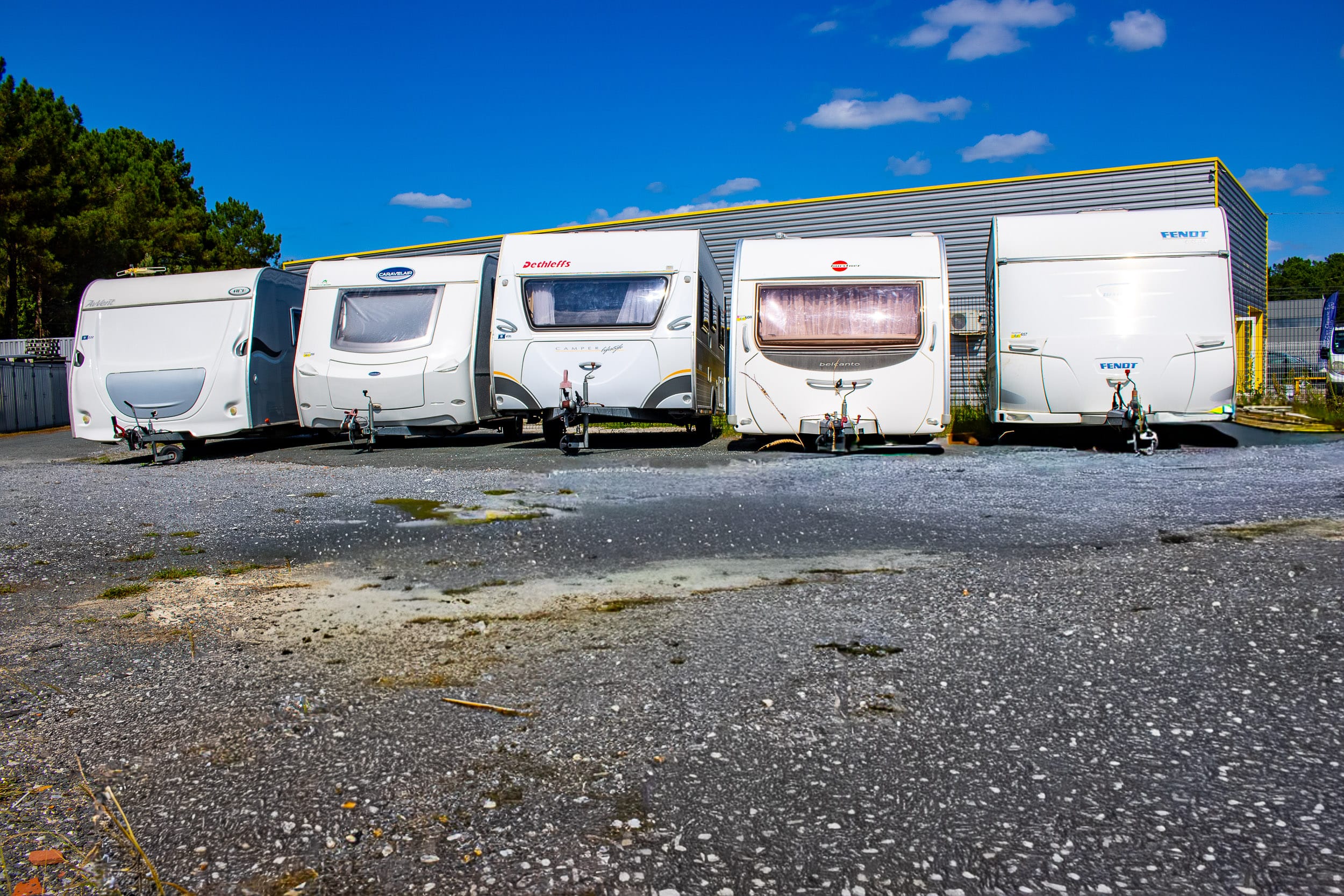 Cinq caravanes blanches (ACE, Caravelair, Dethleffs, Bürstner, Fendt) alignées sur un parking gravier devant un bâtiment moderne.