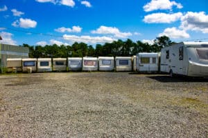 Parking en gravier avec une rangée de caravanes sous un ciel bleu et des nuages. Arbres et entrepôt en arrière-plan. Caravelair visible.