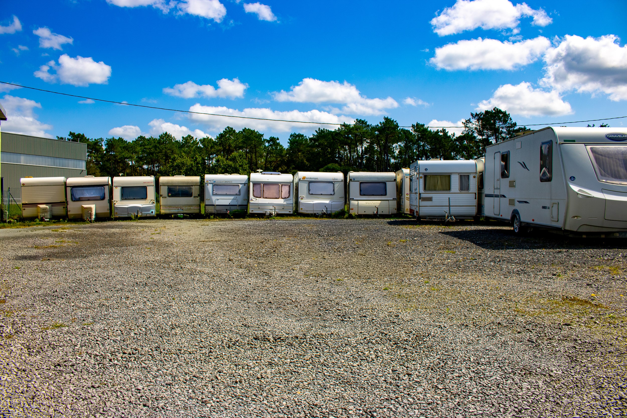 Parking en gravier avec une rangée de caravanes sous un ciel bleu et des nuages. Arbres et entrepôt en arrière-plan. Caravelair visible.