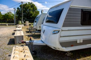 Caravanes Tabbert blanches et autres modèles garées sur un parking extérieur gravillonné. Blocs béton, poteaux et arbres sous ciel bleu.