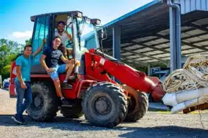 Trois hommes souriants près d'un chargeur frontal rouge avec des bâches en vrac. L'un est dans la cabine, deux autres sur/à côté de l'engin.