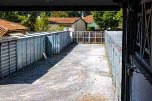 Allée de box de stockage métalliques extérieurs en gravier, bordée de maisons et arbres. Des caméras de surveillance sont visibles à droite.