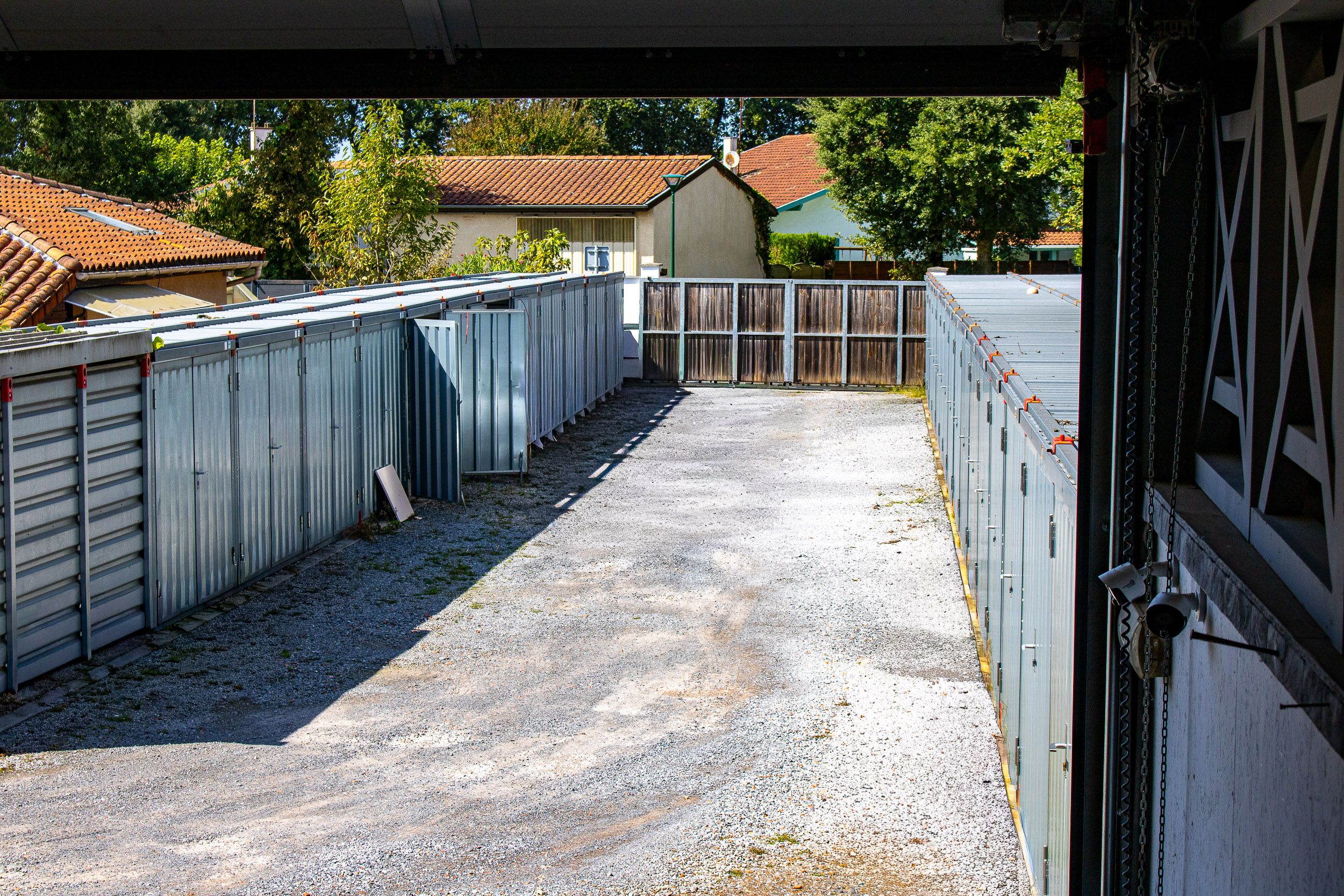 Allée de box de stockage métalliques extérieurs en gravier, bordée de maisons et arbres. Des caméras de surveillance sont visibles à droite.