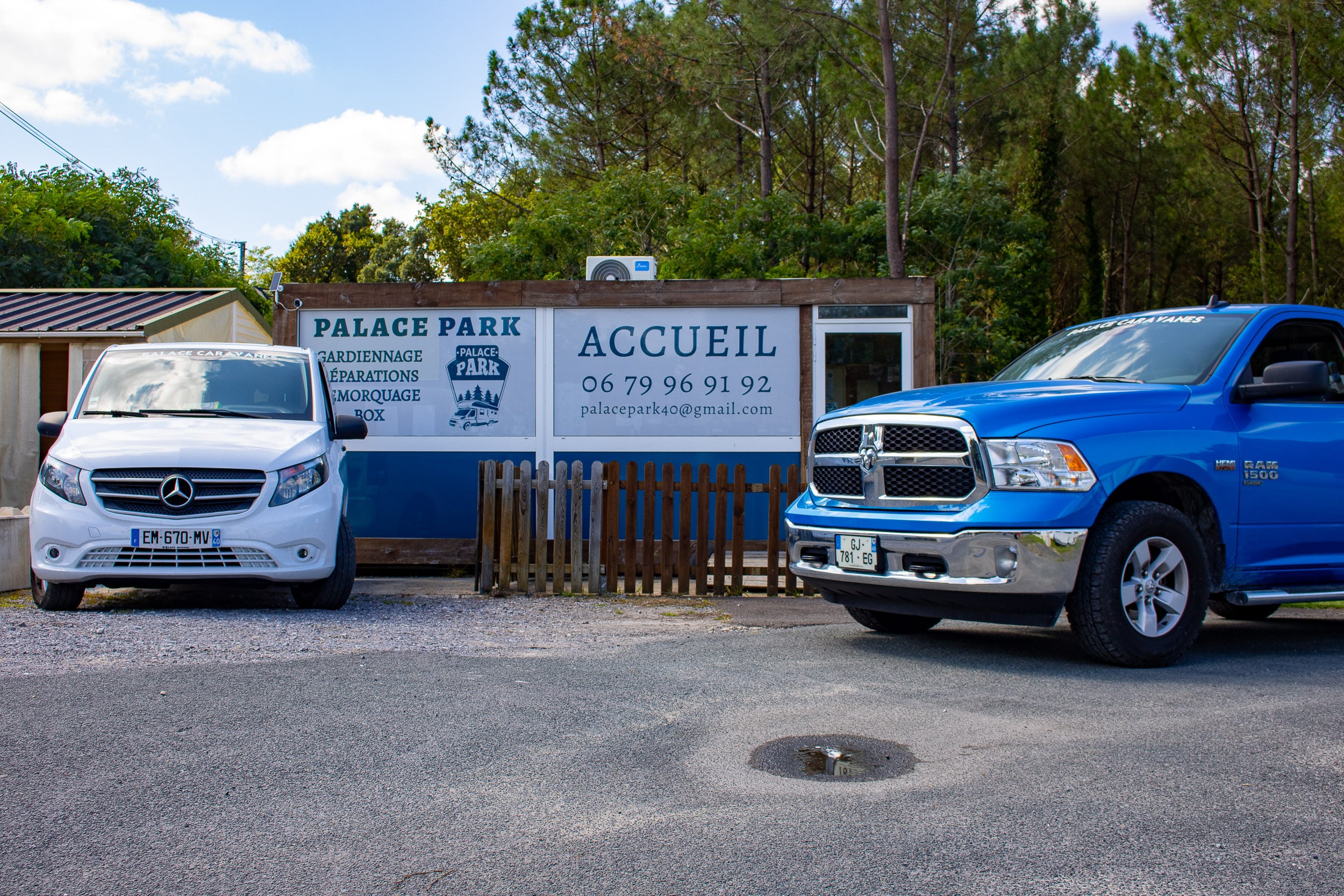 Un van Mercedes blanc et un pick-up RAM bleu devant les panneaux Palace Park affichant accueil, gardiennage, réparations, remorquage et coordonnées de contact.