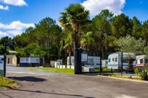 Entrée de Palace Park, un parc de caravanes. Panneaux 'ACCUEIL', services, tél. 06 79 96 92. Palmiers, pins et ciel bleu. Caravanes garées.