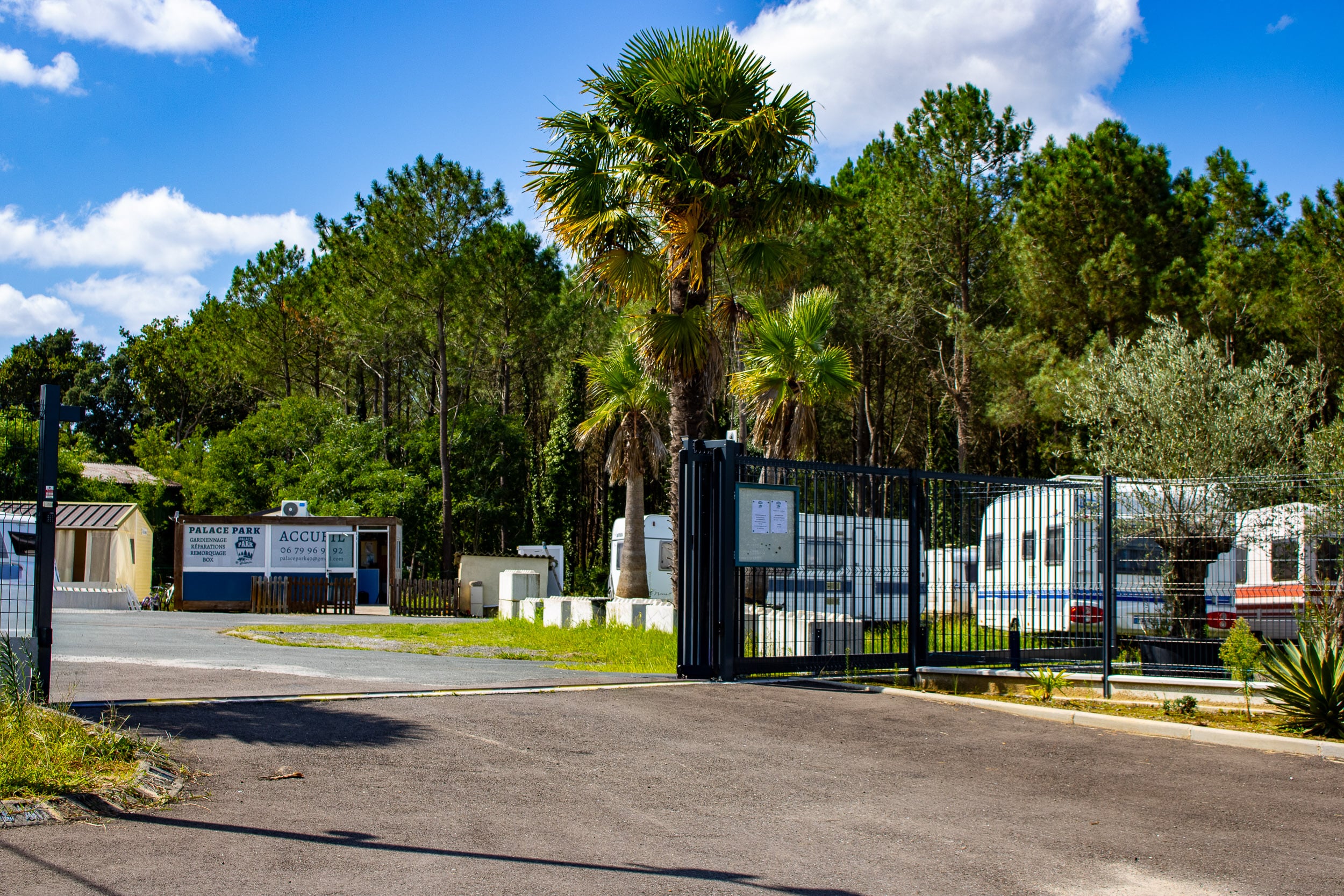 Entrée de Palace Park, un parc de caravanes. Panneaux 'ACCUEIL', services, tél. 06 79 96 92. Palmiers, pins et ciel bleu. Caravanes garées.