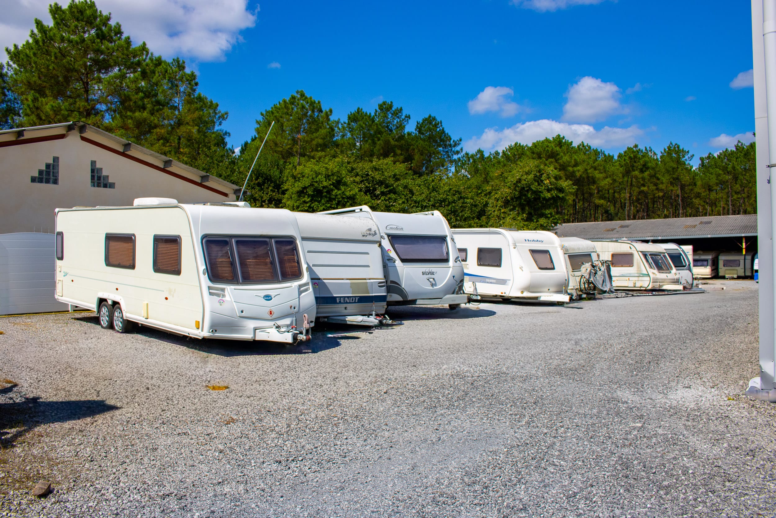 Caravanes blanches de marques Bailey, Fendt et Hobby garées en rang sur un parking extérieur en gravier sous un ciel bleu clair.