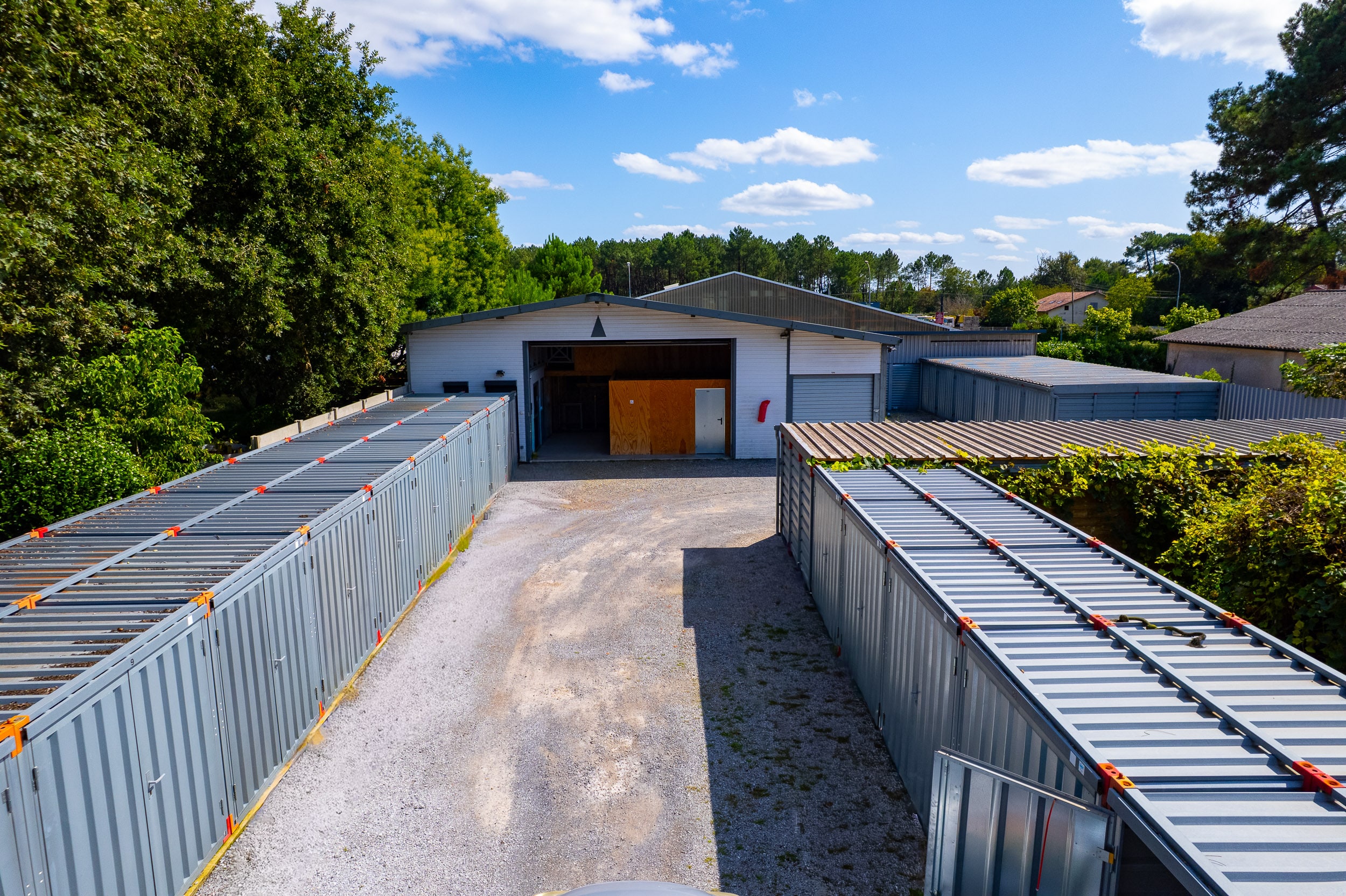 Installation de self-stockage extérieur: entrepôt central et rangées de boxes individuels en métal gris, sous ciel bleu, entourée d'arbres.