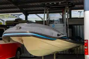 Bateau semi-rigide gris et blanc avec bande bleue, bâché, stocké sous un abri. Voiture rouge et autre embarcation derrière.