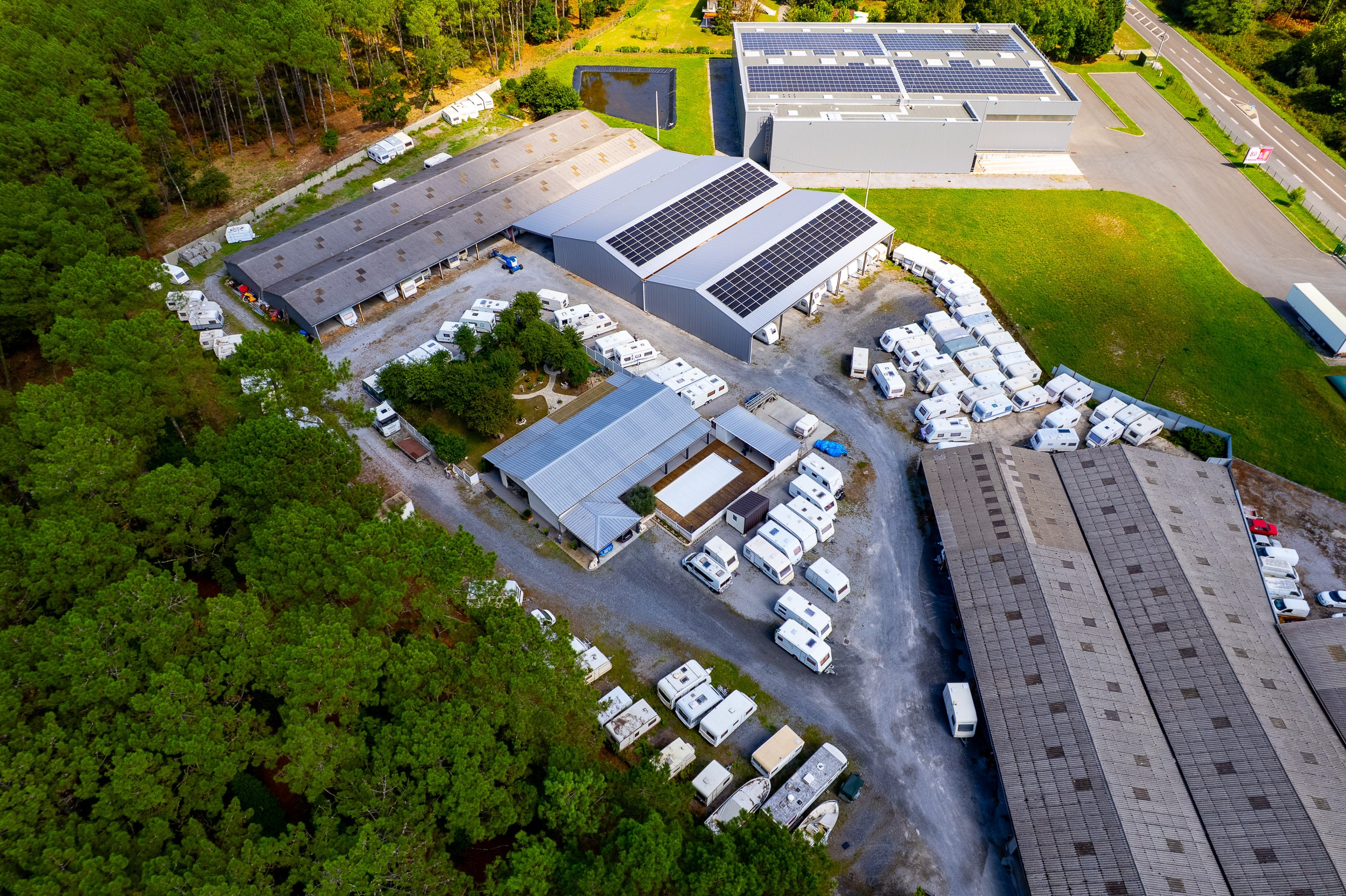 Vue aérienne d'un complexe industriel : bâtiments à panneaux solaires, stockage de caravanes/camping-cars, forêt et route (panneau '80').