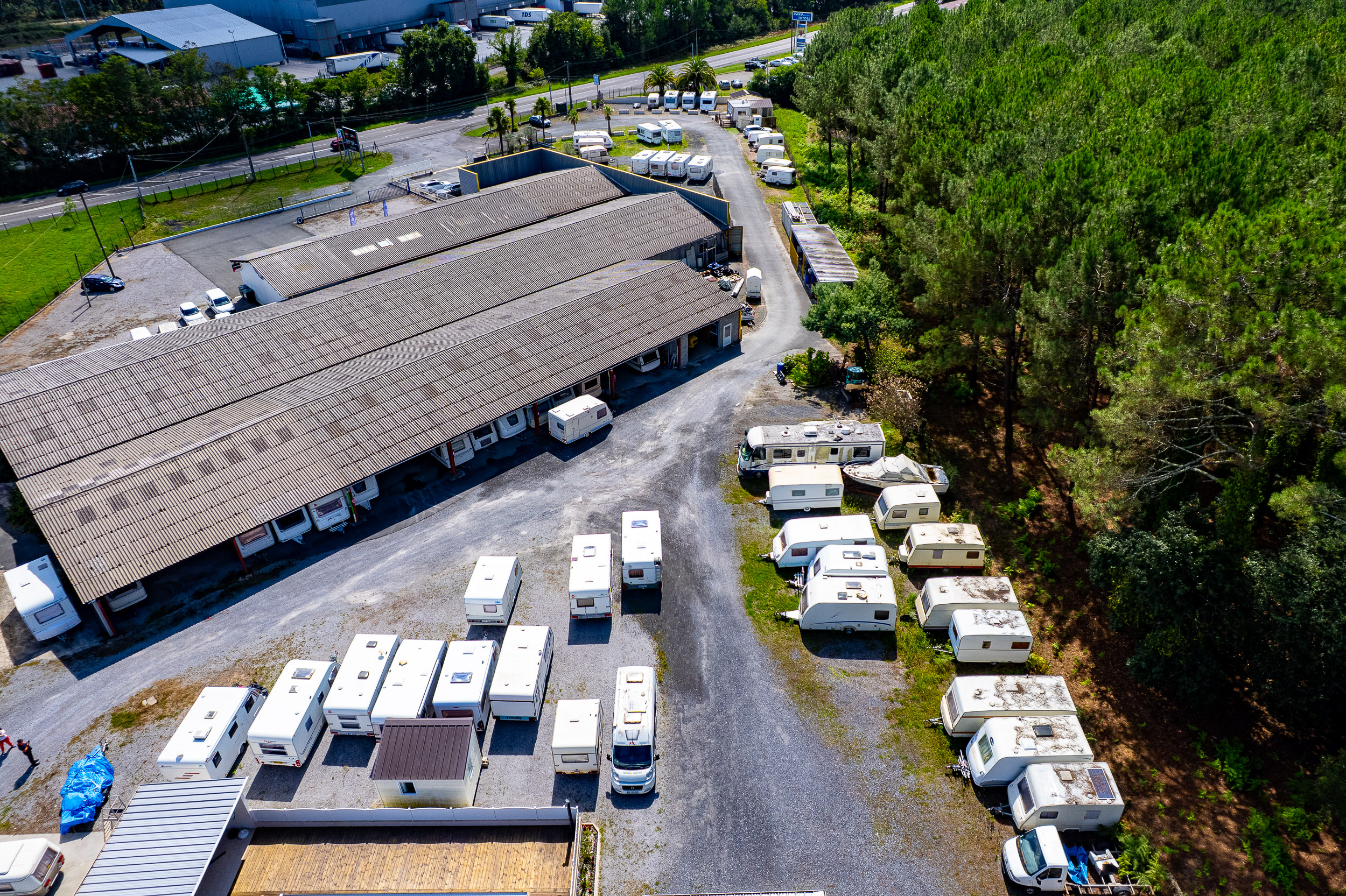 Vue aérienne d'un parc de stockage de caravanes et camping-cars blancs, stationnés en plein air et sous abris. Bâtiments et forêt en arrière-plan.