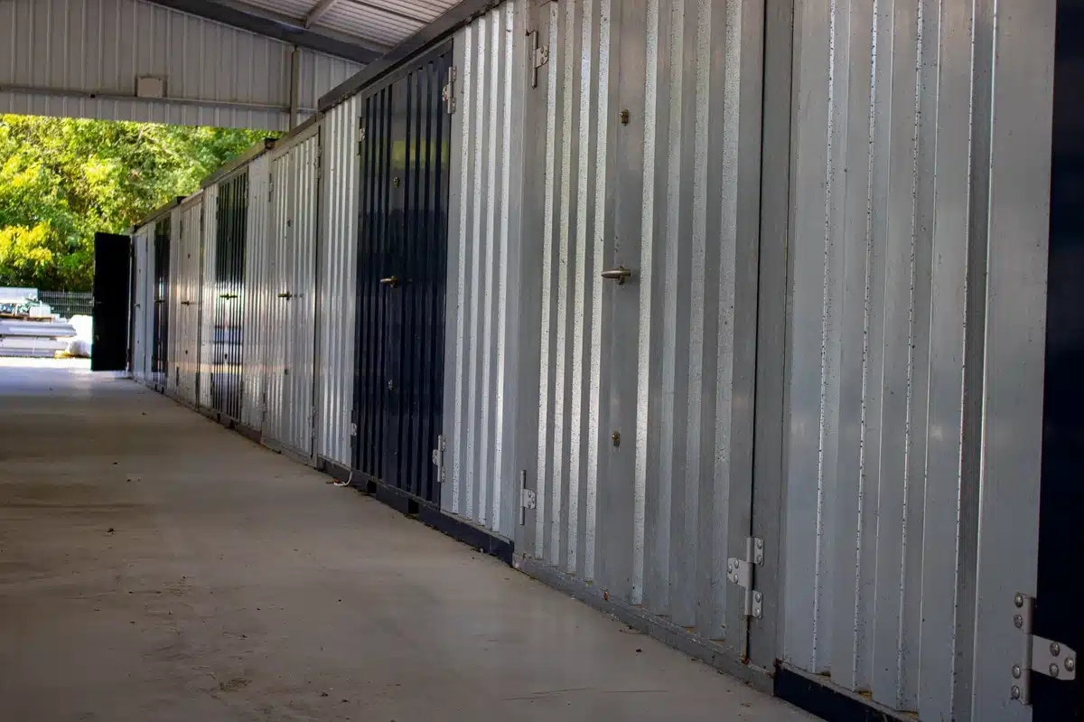 Rangée d'unités de stockage en métal galvanisé et noir sous un abri. Une porte est ouverte, montrant des arbres en arrière-plan.