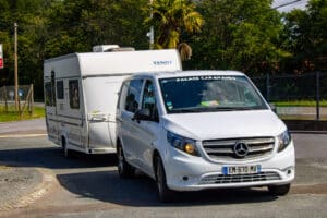 Van Mercedes blanc "PALACE CARAVANES" tractant une caravane Fendt blanche sur une route ensoleillée, entourée d'arbres.