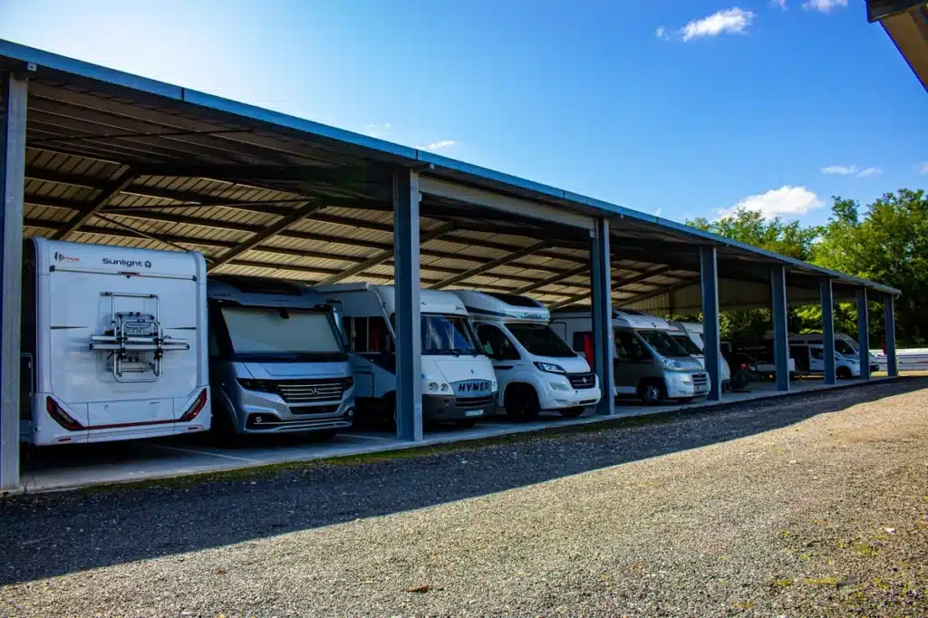 Plusieurs camping-cars de marques Sunlight, Hymer et Challenger sont garés sous un grand abri métallique extérieur, sur un parking en gravier, sous un ciel bleu ensoleillé.
