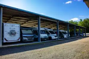 Plusieurs camping-cars de marques Sunlight, Hymer et Challenger sont garés sous un grand abri métallique extérieur, sur un parking en gravier, sous un ciel bleu ensoleillé.