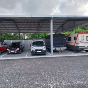 Abri de parking métallique pour véhicules : SUV bordeaux, fourgonnette blanche, remorque bâchée et camping-car garés sur gravier.