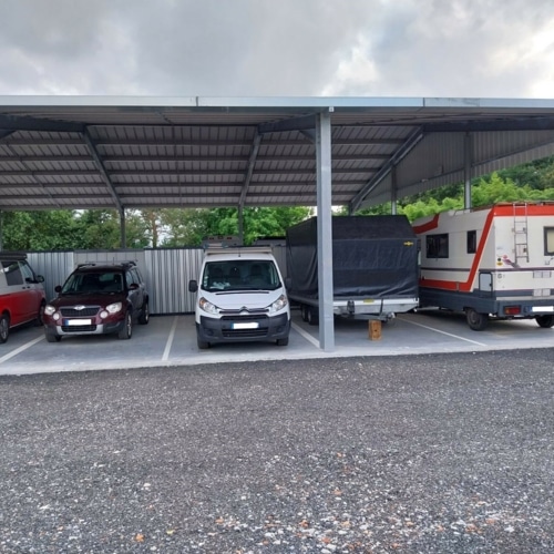 Abri de parking métallique pour véhicules : SUV bordeaux, fourgonnette blanche, remorque bâchée et camping-car garés sur gravier.