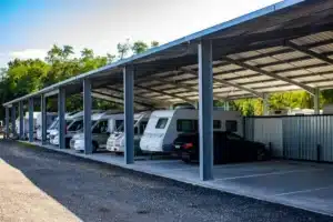 Long abri métallique protégeant des camping-cars, caravanes et une voiture noire garés en rangée sur un parking extérieur. Arbres et ciel bleu.